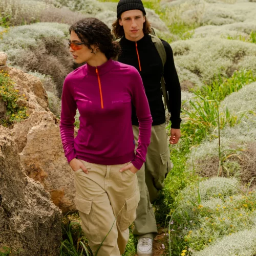 Dwóch wędrowców idzie przez zielony, skalisty krajobraz. Pierwszy z nich ma na sobie koszulkę Bluza z wełny merino trekkingowa damska fuksja ze spodniami w kolorze khaki i okularami przeciwsłonecznymi; drugi ma na sobie czarną koszulę z długim rękawem, spodnie w kolorze khaki i czarną czapkę.
