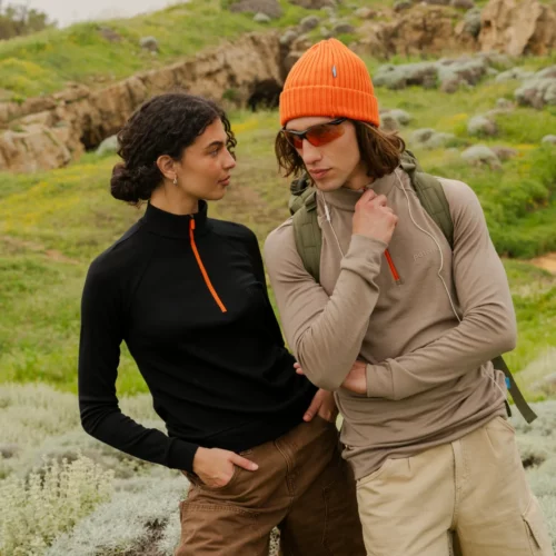 Dwie osoby stoją na trawiastym wzgórzu. Jedna z nich ma na sobie fuksjową koszulkę Bluza z wełny merino trekkingowa damska w połączeniu z brązowymi szortami. Druga ma na sobie brązową koszulkę z długim rękawem, pomarańczową czapkę, okulary przeciwsłoneczne, beżowe szorty i plecak.