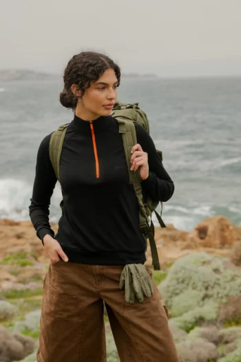 Kobieta stoi nad oceanem w fuksjowej bluzie z wełny merino trekkingowej damskiej, brązowych spodniach, z zielonym plecakiem i zielonymi rękawiczkami w kieszeni. W tle widoczna jest skalista linia brzegowa i woda.