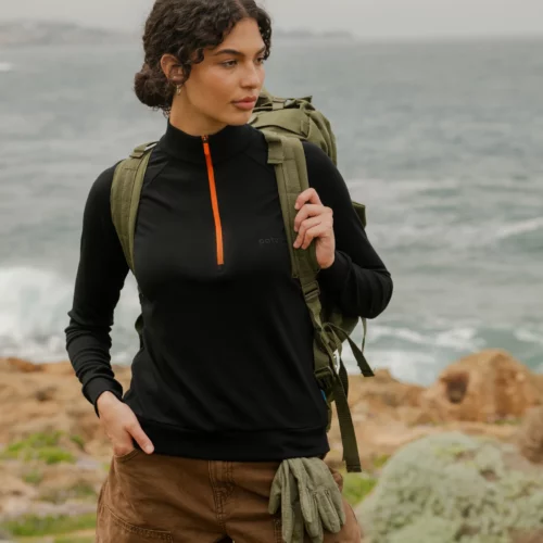 Kobieta stoi nad oceanem w fuksjowej bluzie z wełny merino trekkingowej damskiej, brązowych spodniach, z zielonym plecakiem i zielonymi rękawiczkami w kieszeni. W tle widoczna jest skalista linia brzegowa i woda.