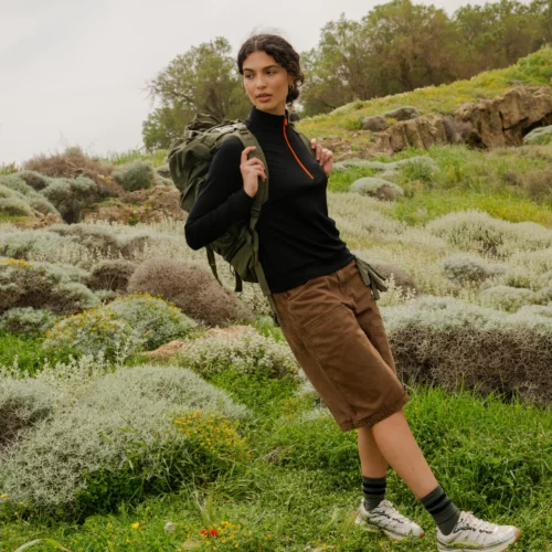 Osoba stoi na zewnątrz na trawiastym terenie z krzewami, ubrana w koszulkę "Bluza z wełny merino trekkingowa damska fuksja", brązowe szorty, trampki i zielony plecak, patrząc w bok.