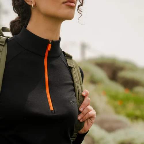 Kobieta z kręconymi włosami nosi Bluzę z wełny merino trekkingową damską fuksję i niesie plecak, stojąc pewnie na zewnątrz, z rozmytą zielenią i polnymi kwiatami w tle.