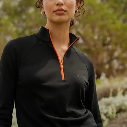 Osoba z kręconymi włosami stoi na zewnątrz w delikatnym świetle słonecznym, ubrana w sweter Bluza z wełny merino trekkingowa damska fuksja. W tle widoczne są zielone liście.