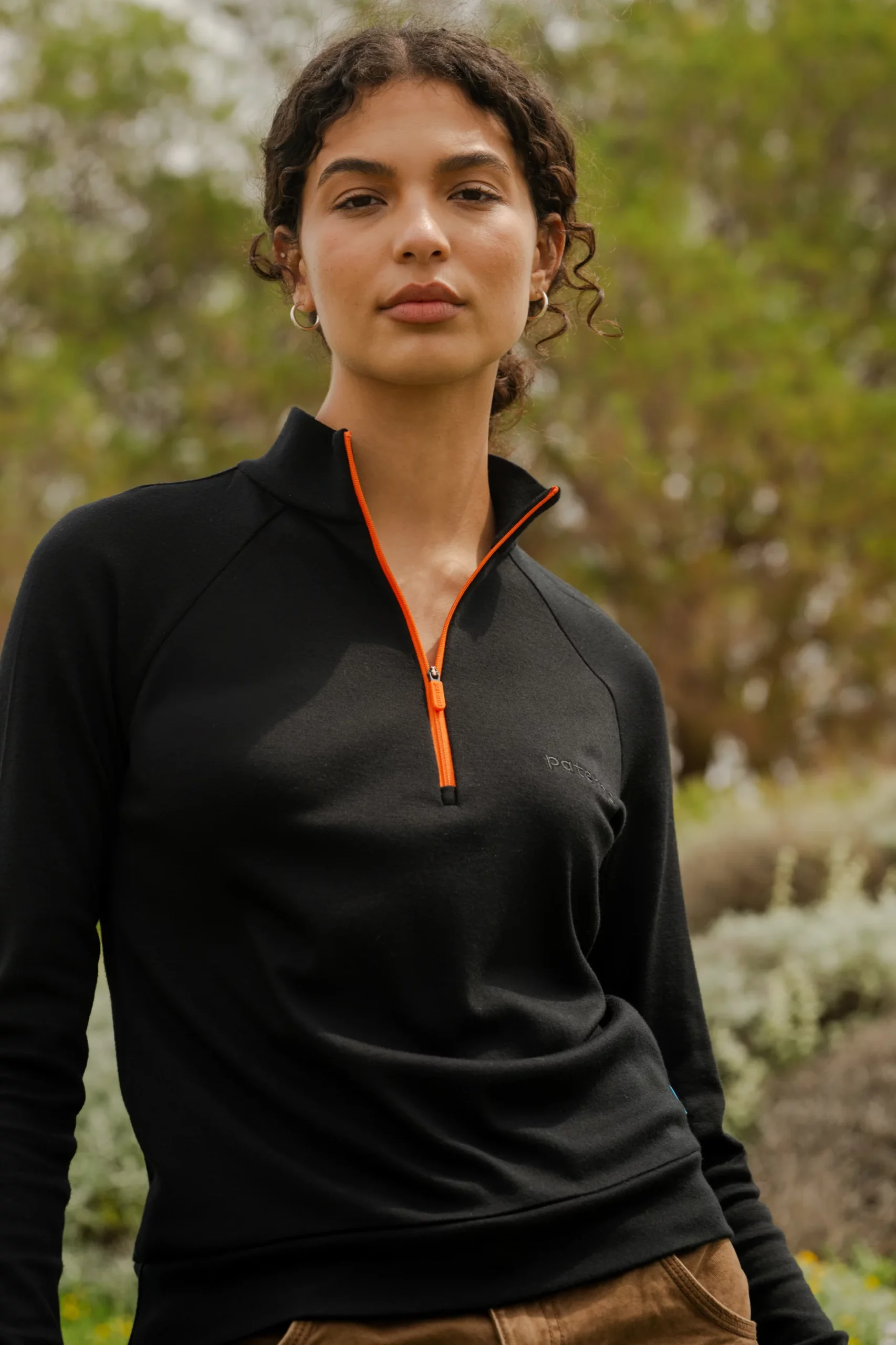 Osoba z kręconymi włosami stoi na zewnątrz w delikatnym świetle słonecznym, ubrana w sweter Bluza z wełny merino trekkingowa damska fuksja. W tle widoczne są zielone liście.