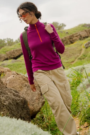 Osoba ubrana w koszulkę "Bluza z wełny merino trekkingowa damska fuksja", spodnie cargo w kolorze khaki, okulary przeciwsłoneczne i plecak stoi zamyślona na trawiastym, skalistym wzgórzu na zewnątrz.