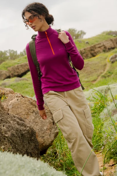 Osoba ubrana w koszulkę "Bluza z wełny merino trekkingowa damska fuksja", spodnie cargo w kolorze khaki, okulary przeciwsłoneczne i plecak stoi zamyślona na trawiastym, skalistym wzgórzu na zewnątrz.
