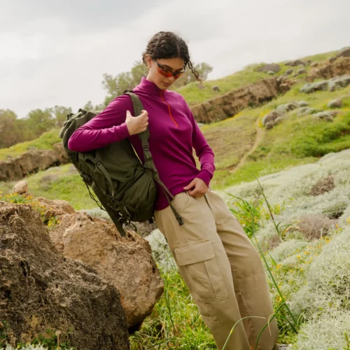 Osoba ubrana w Bluzę z wełny merino trekkingową damską fuksję, spodnie cargo w kolorze khaki i okulary przeciwsłoneczne stoi na skałach wśród zielonej trawy, niosąc duży zielony plecak i patrząc w dół.
