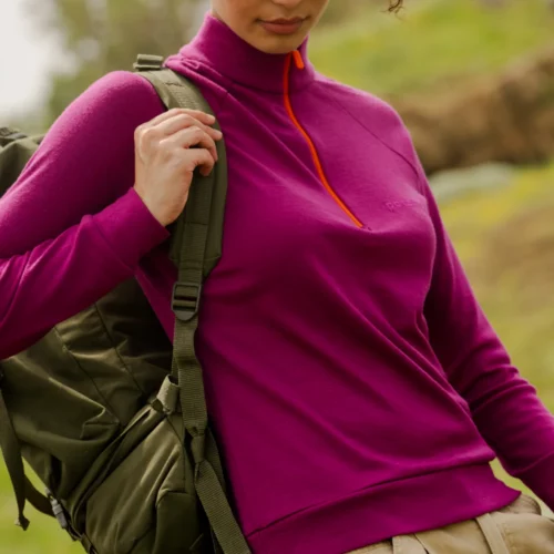 Kobieta ubrana w koszulkę "Bluza z wełny merino trekkingowa damska fuksja" stoi na zewnątrz na trawiastym terenie, w połączeniu ze spodniami khaki, czerwonymi okularami przeciwsłonecznymi i zielonym plecakiem na tle rozmytych wzgórz.