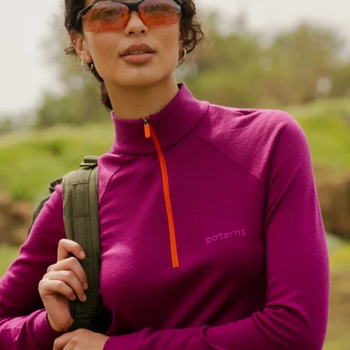 Osoba na zewnątrz ma na sobie "Bluzę z wełny merino trekkingową damską fuksję" we wzory, okulary przeciwsłoneczne w pomarańczowym odcieniu i niesie zielony plecak. Tło jest trawiaste i rozmyte.