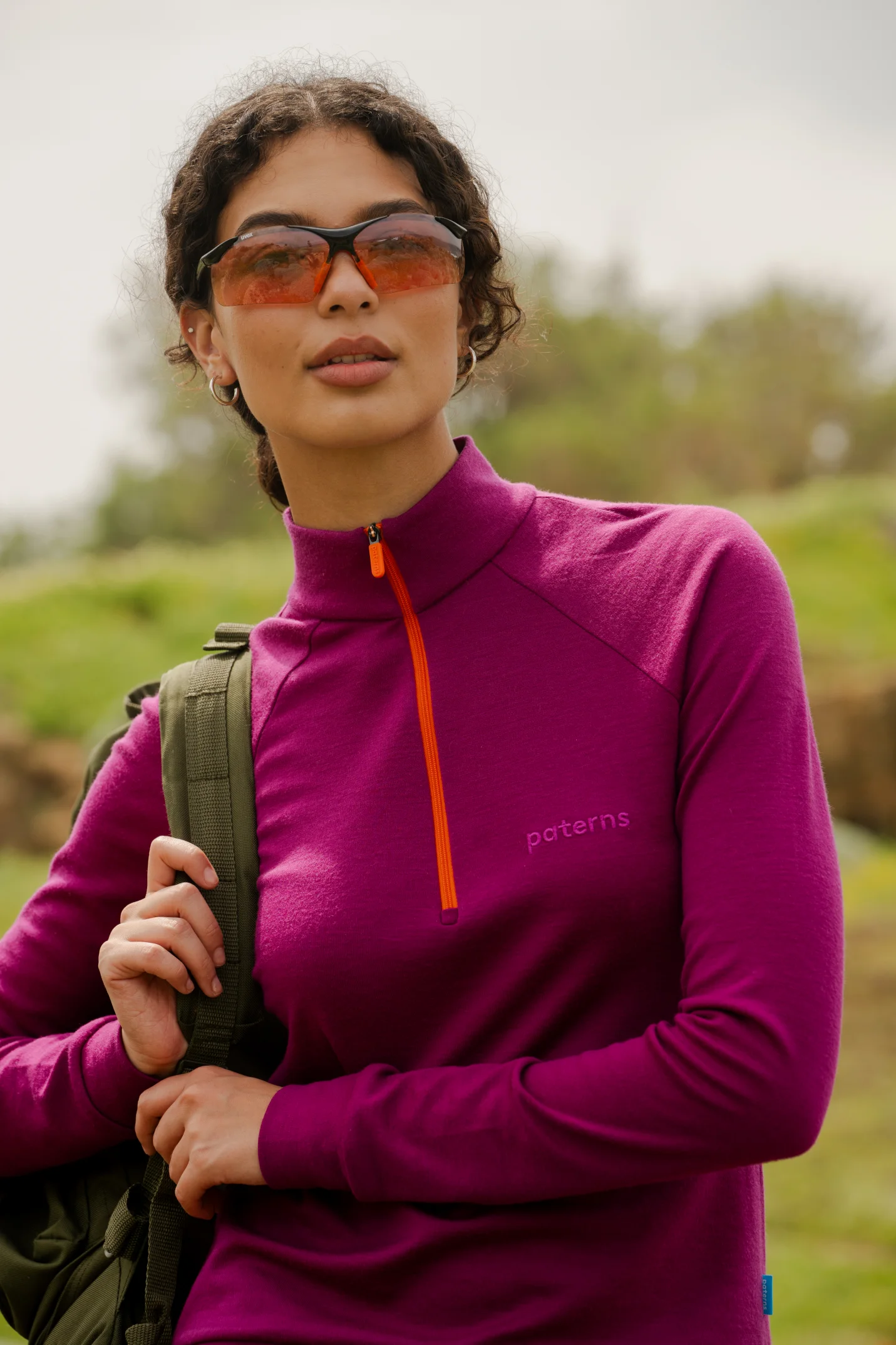 Osoba na zewnątrz ma na sobie "Bluzę z wełny merino trekkingową damską fuksję" we wzory, okulary przeciwsłoneczne w pomarańczowym odcieniu i niesie zielony plecak. Tło jest trawiaste i rozmyte.