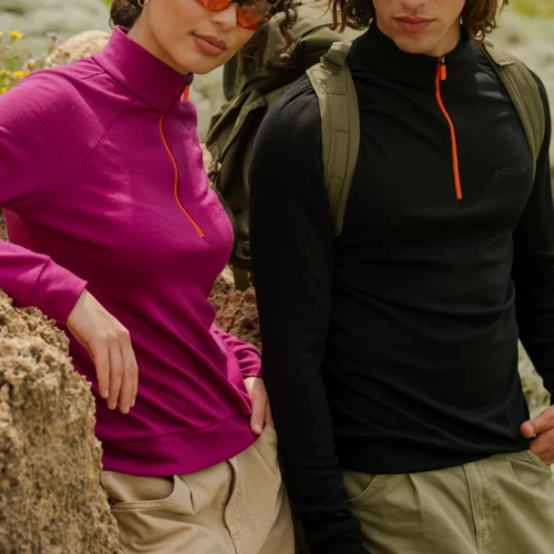 Dwie osoby na zewnątrz opierają się o skałę. Osoba po lewej ma na sobie trekkingową damską Bluzę z wełny merino w kolorze fuksji, brązowe spodnie i pomarańczowe okulary przeciwsłoneczne, natomiast osoba po prawej ma na sobie czarny top zapinany na zamek, oliwkowe spodnie i plecak.