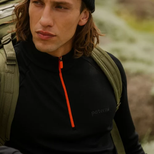 Kobieta ubrana w Bluzę z wełny merino trekkingową damską fuksja siedzi na zewnątrz z poważnym wyrazem twarzy, otoczona rozmytą zielenią i skałami.