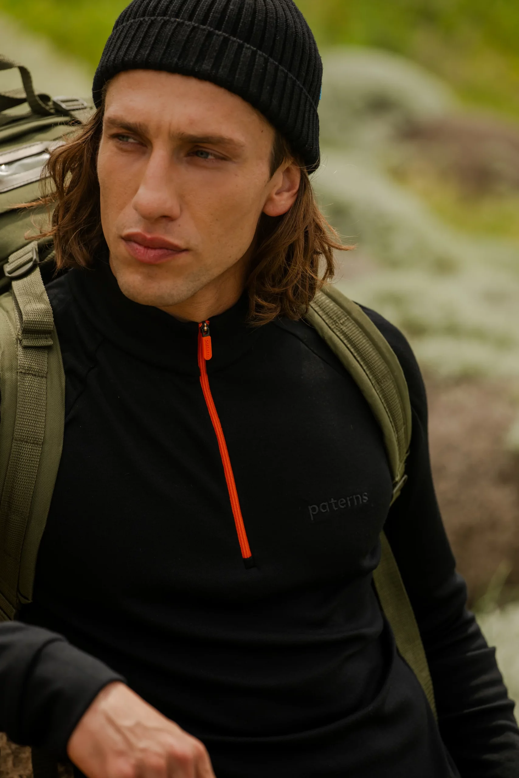 Kobieta ubrana w Bluzę z wełny merino trekkingową damską fuksja siedzi na zewnątrz z poważnym wyrazem twarzy, otoczona rozmytą zielenią i skałami.