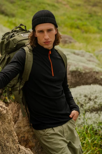 Osoba ubrana w Bluza z wełny merino trekkingowa damska fuksja stoi na zewnątrz na skalistym terenie z zieloną trawą w tle, w połączeniu z czarną czapką, spodniami khaki i plecakiem.