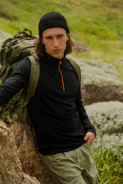 Osoba ubrana w Bluza z wełny merino trekkingowa damska fuksja stoi na zewnątrz na skalistym terenie z zieloną trawą w tle, w połączeniu z czarną czapką, spodniami khaki i plecakiem.