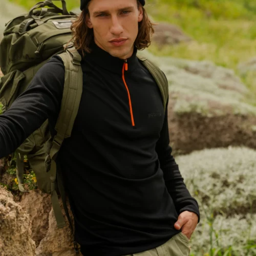 Osoba ubrana w Bluza z wełny merino trekkingowa damska fuksja stoi na zewnątrz na skalistym terenie z zieloną trawą w tle, w połączeniu z czarną czapką, spodniami khaki i plecakiem.