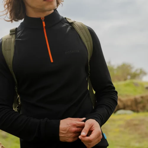 Osoba nosząca Bluzę z wełny merino trekkingową damską fuksja stoi na zewnątrz wśród zieleni pod pochmurnym niebem, patrząc w zamyśleniu w bok.