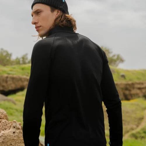 Kobieta ubrana w bluzę trekkingową z wełny merynosów w kolorze fuksji stoi na zewnątrz, spoglądając przez ramię. Zielona trawa i duże skały pojawiają się w tle pod zachmurzonym niebem.