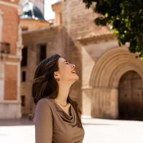 Kobieta o długich brązowych włosach, nosząca Koszulkę z wełny merino prosta damska mokka, uśmiecha się i spogląda w górę, stojąc na zewnątrz na oświetlonym słońcem placu z zabytkowymi kamiennymi budynkami i łukowatymi drzwiami za nią.