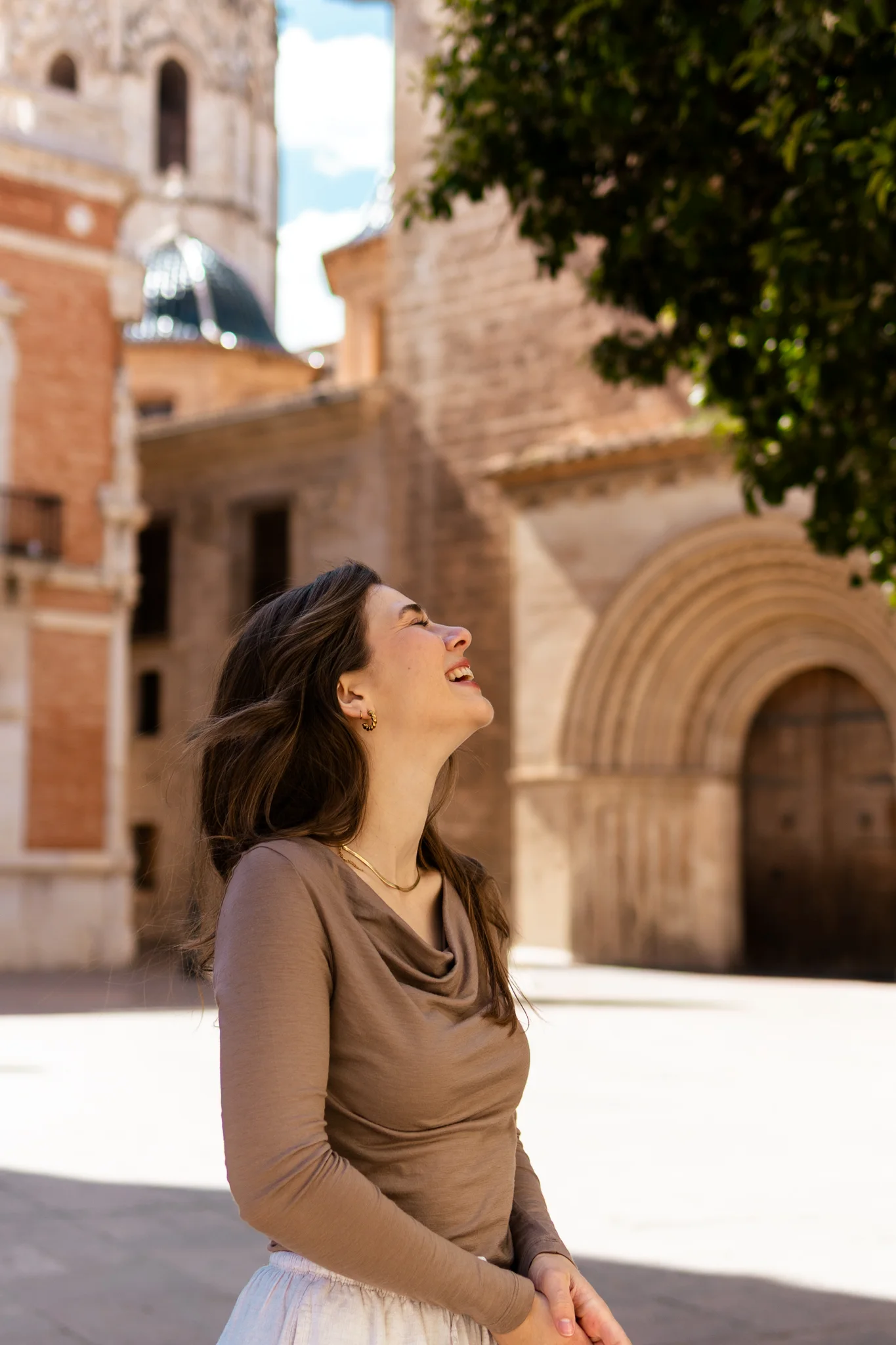 Kobieta o długich brązowych włosach, nosząca Koszulkę z wełny merino prosta damska mokka, uśmiecha się i spogląda w górę, stojąc na zewnątrz na oświetlonym słońcem placu z zabytkowymi kamiennymi budynkami i łukowatymi drzwiami za nią.