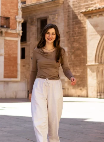 Młoda kobieta z długimi brązowymi włosami, ubrana w koszulkę z wełny merino prosta damska mokka i białe spodnie, uśmiecha się, wychodząc na słoneczny dziedziniec z zabytkowymi kamiennymi budynkami w tle.