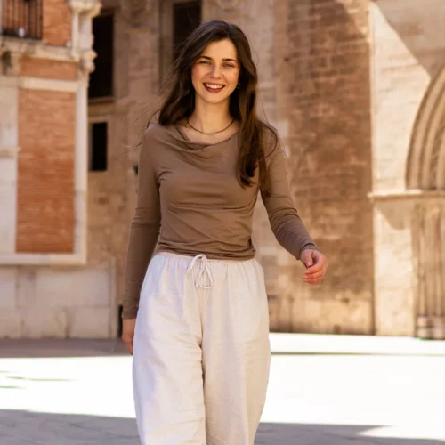 Młoda kobieta z długimi brązowymi włosami, ubrana w koszulkę z wełny merino prosta damska mokka i białe spodnie, uśmiecha się, wychodząc na słoneczny dziedziniec z zabytkowymi kamiennymi budynkami w tle.