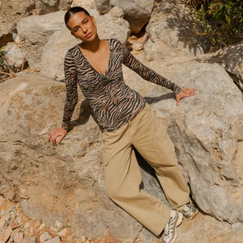 Kobieta ubrana w Koszulkę z wełny merino z rękawem 3/4 mokka siedzi na dużych skałach na zewnątrz, z zamkniętymi oczami i głową odchyloną do tyłu, otoczona naturalnym kamieniem i zielenią.