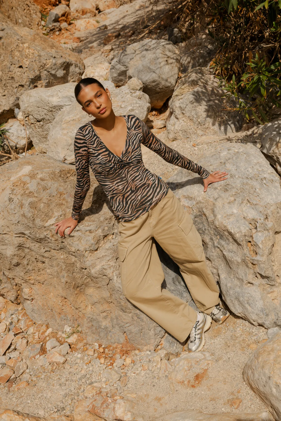 Kobieta ubrana w Koszulkę z wełny merino z rękawem 3/4 mokka siedzi na dużych skałach na zewnątrz, z zamkniętymi oczami i głową odchyloną do tyłu, otoczona naturalnym kamieniem i zielenią.