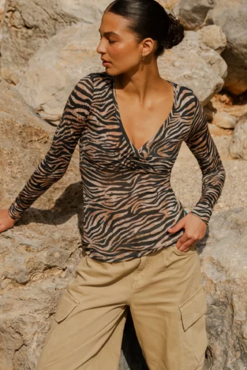 Kobieta z ciemnymi włosami spiętymi w kok nosi "Koszulkę z wełny merynos z rękawem 3/4 mokka", pozując na skalistym tle i spoglądając w lewo.