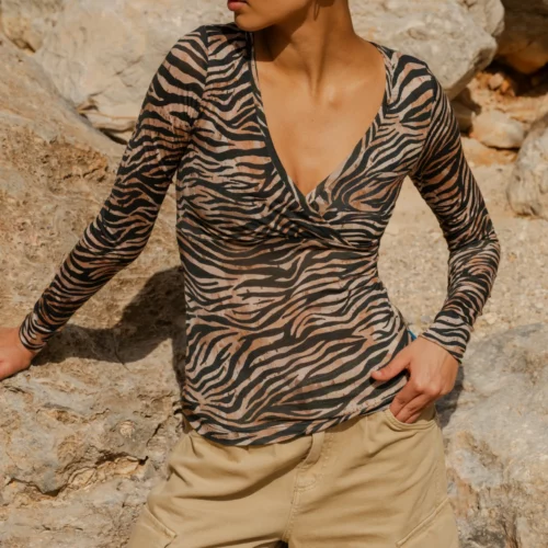 Kobieta z ciemnymi włosami spiętymi w kok nosi "Koszulkę z wełny merynos z rękawem 3/4 mokka", pozując na skalistym tle i spoglądając w lewo.