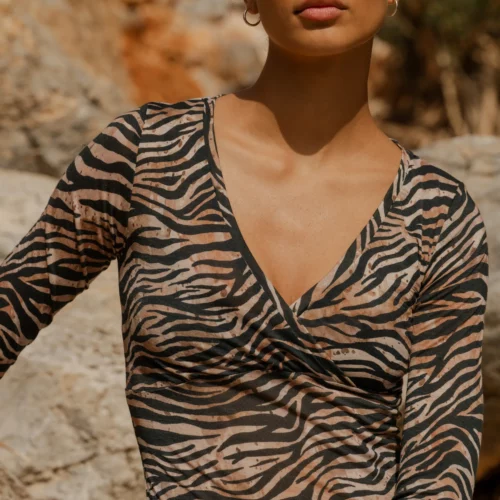 Kobieta z zaczesanymi do tyłu włosami nosi Koszulkę z wełny merino z rękawem 3/4 mokka, pozując pewnie w plenerze przed dużymi skałami i patrząc w kamerę.