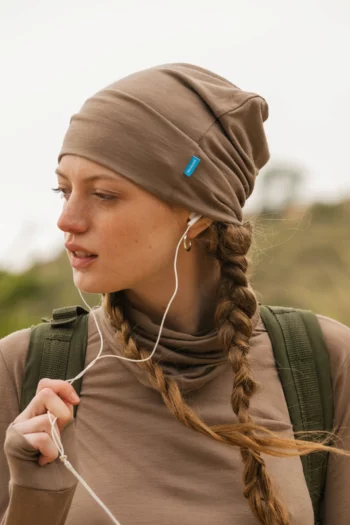 Młoda kobieta z dwoma warkoczami ma na sobie czapkę z wełny merynosów w kolorze mokka (Czapka z wełny merynosów cienka mokka) i pasujący top, słuchając muzyki przez białe słuchawki. Ma zielony plecak i stoi na zewnątrz na rozmytym naturalnym tle.