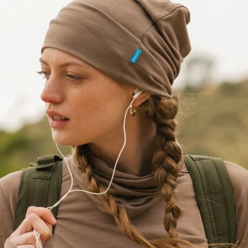 Młoda kobieta z dwoma warkoczami ma na sobie czapkę z wełny merynosów w kolorze mokka (Czapka z wełny merynosów cienka mokka) i pasujący top, słuchając muzyki przez białe słuchawki. Ma zielony plecak i stoi na zewnątrz na rozmytym naturalnym tle.