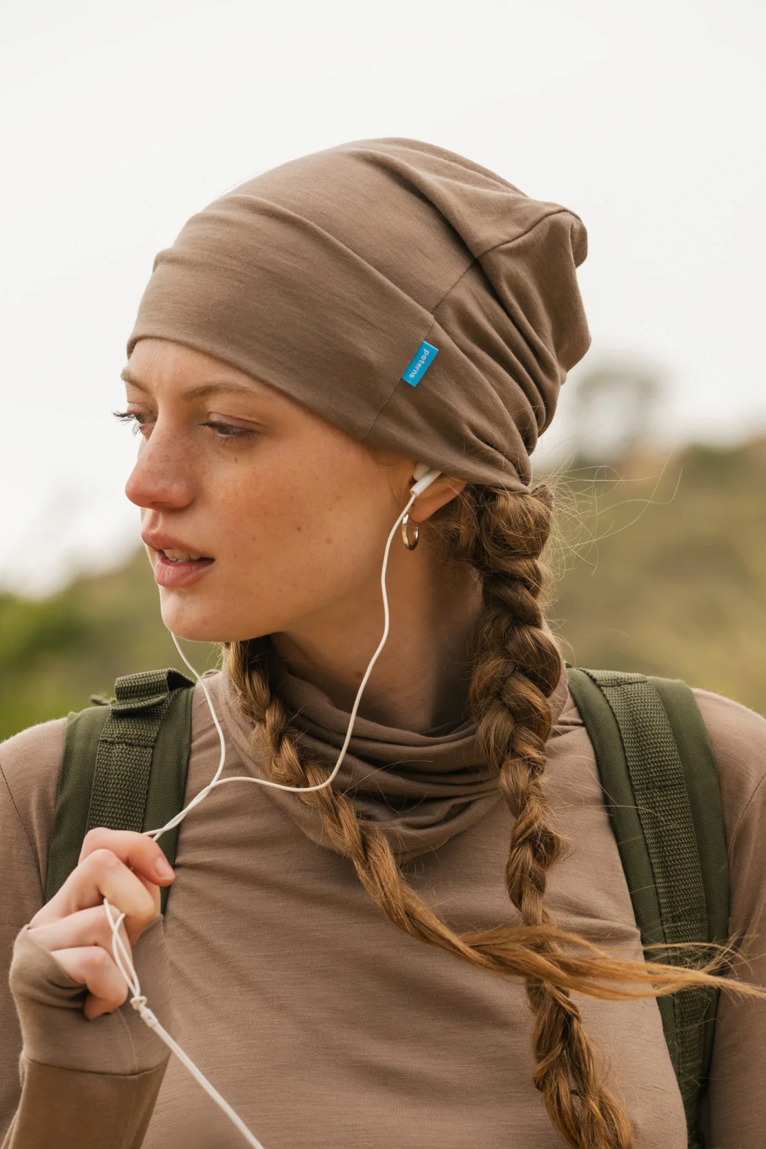 Młoda kobieta z dwoma warkoczami ma na sobie czapkę z wełny merynosów w kolorze mokka (Czapka z wełny merynosów cienka mokka) i pasujący top, słuchając muzyki przez białe słuchawki. Ma zielony plecak i stoi na zewnątrz na rozmytym naturalnym tle.