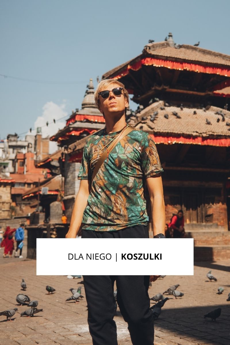 Mężczyzna w okularach przeciwsłonecznych i wzorzystej koszulce stoi na oświetlonym słońcem placu z zabytkowymi budynkami i gołębiami. Nakładka z tekstem DLA NIEGO | KOSZULKI.