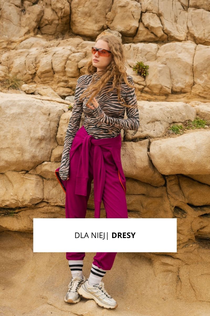 Kobieta w okularach przeciwsłonecznych ma na sobie top z nadrukiem zebry, fioletowe spodnie dresowe i srebrne trampki, pozując na tle skalistego terenu. Nakładka tekstowa brzmi "DLA NIEJ | DRESY".