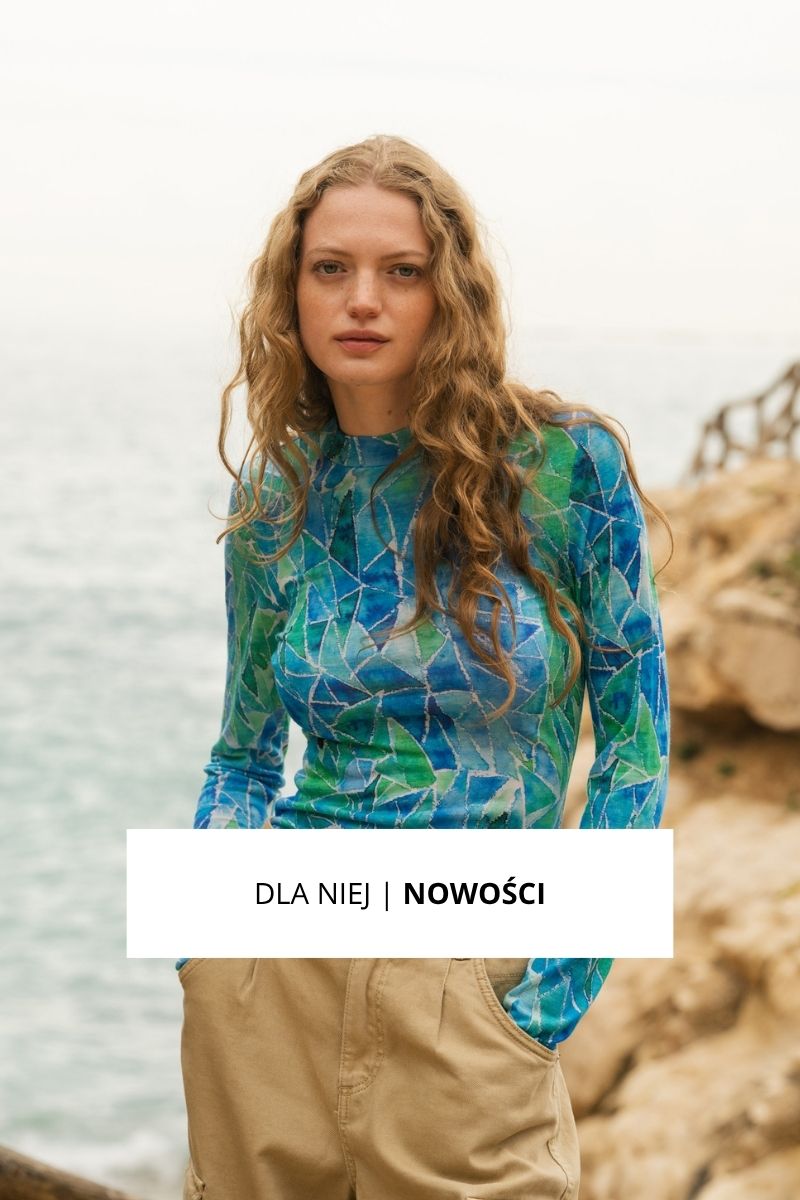 Kobieta z długimi falowanymi włosami stoi nad morzem, ubrana w niebiesko-zieloną wzorzystą koszulę z długim rękawem i beżowe spodnie. Tekst na obrazie brzmi DLA NIEJ | NOWOŚCI.