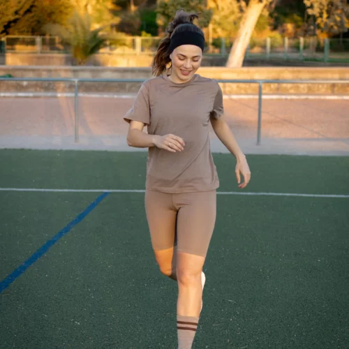 Kobieta ubrana w koszulkę z wełny merino prosta damska mokka biega i uśmiecha się na zielonym boisku sportowym, w tle widać drzewa i ogrodzenie.