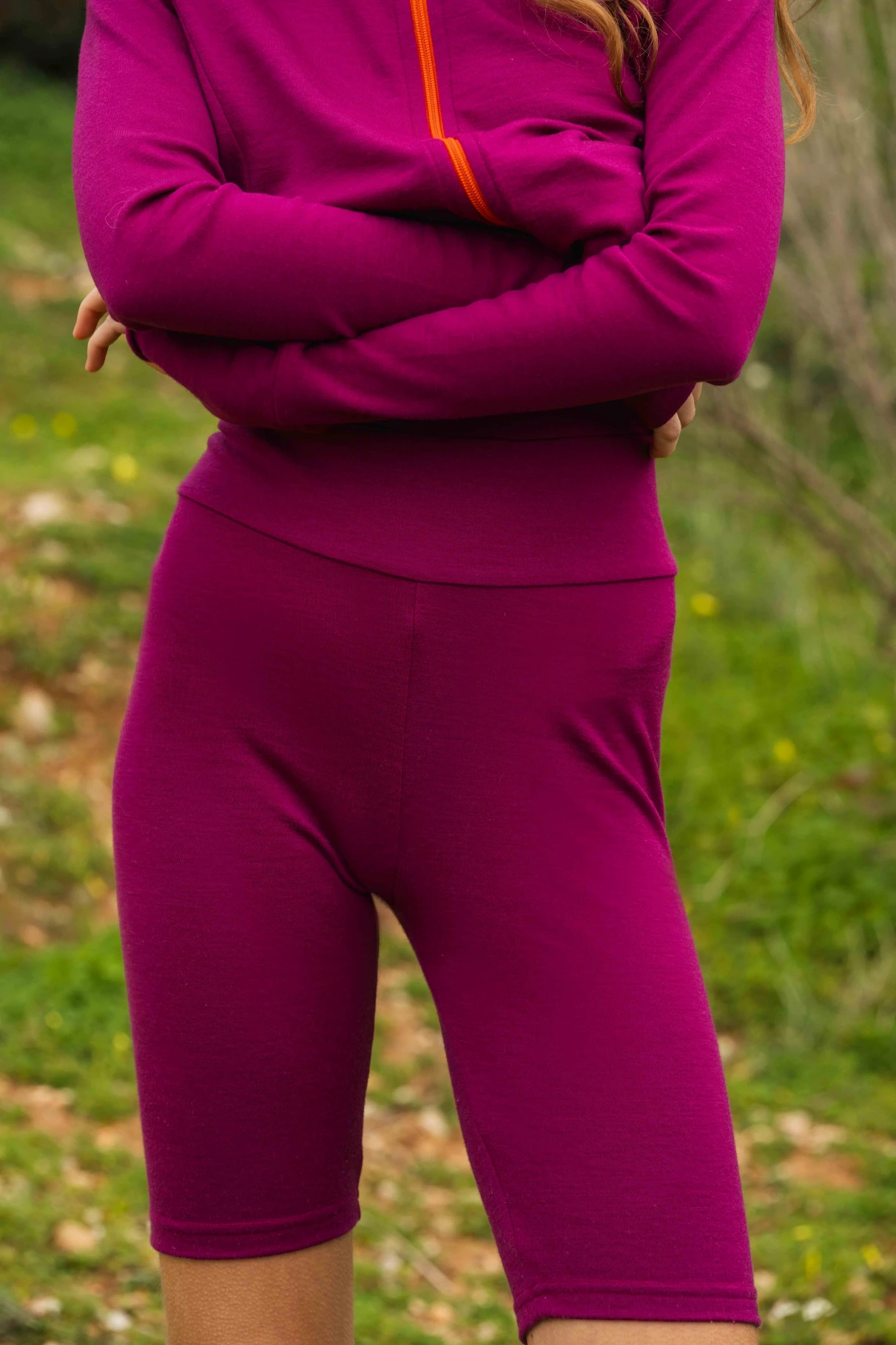 Osoba stoi na zewnątrz ze skrzyżowanymi ramionami, ubrana w Kolarki z wełny merino damskie malinowe. Trawiaste tło jest rozmyte, a twarz nie jest widoczna.