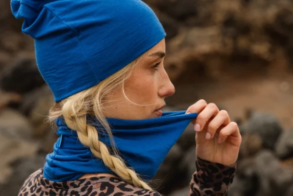 Kobieta z blond warkoczami nosi niebieską czapkę i pasujący ochraniacz na szyję, trzymając ochraniacz przy twarzy. Ubrana jest w bluzkę w panterkę i stoi na zewnątrz w pobliżu skał.