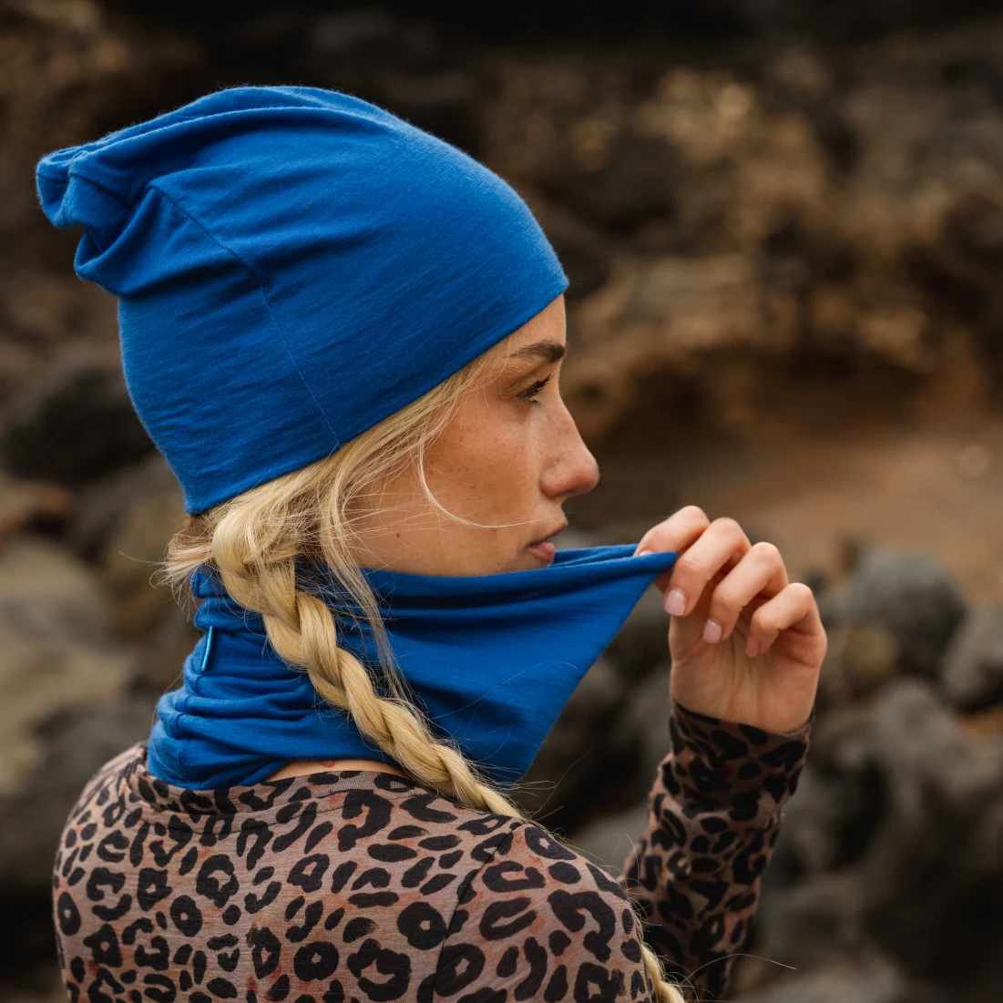 Kobieta z blond warkoczami nosi niebieską czapkę i pasujący ochraniacz na szyję, trzymając ochraniacz przy twarzy. Ubrana jest w bluzkę w panterkę i stoi na zewnątrz w pobliżu skał.