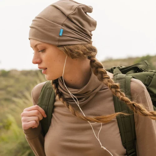 Osoba z włosami splecionymi w warkocz ma na sobie opaloną bluzkę z długim rękawem i dopasowaną Czapka z wełny merino cienka mokka, niosąc zielony plecak i słuchając słuchawek na trawiastym terenie zewnętrznym.