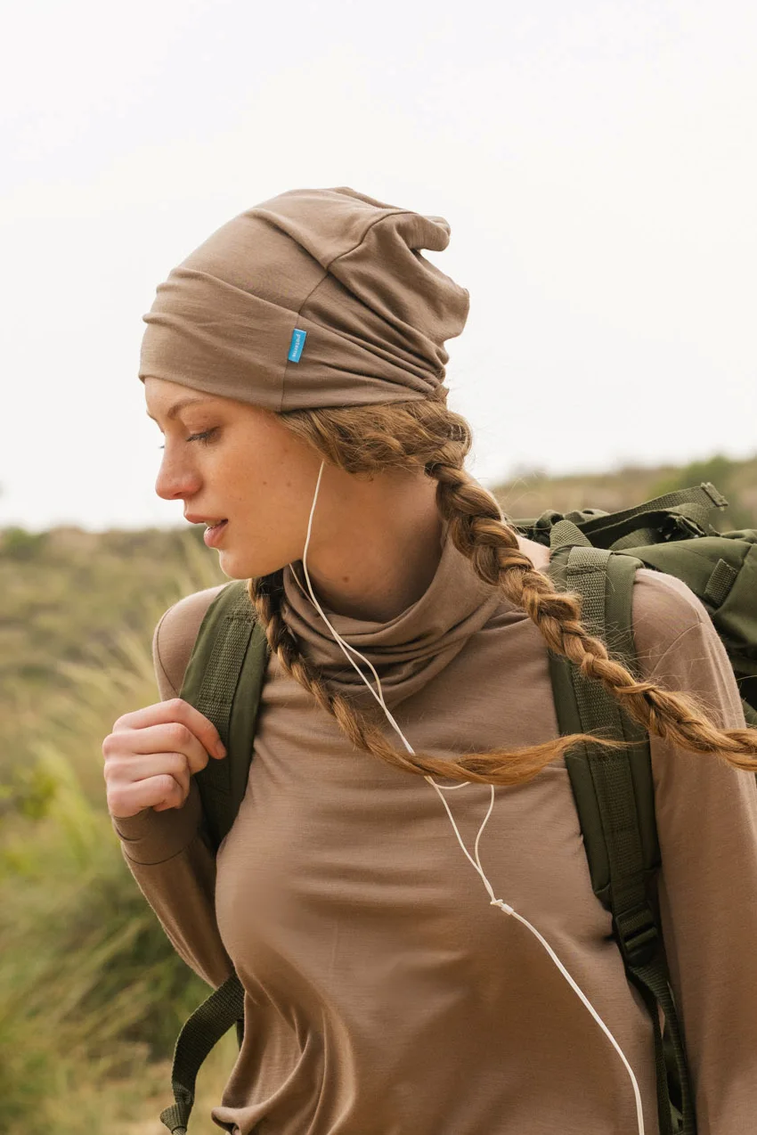 Osoba z włosami splecionymi w warkocz ma na sobie opaloną bluzkę z długim rękawem i dopasowaną Czapka z wełny merino cienka mokka, niosąc zielony plecak i słuchając słuchawek na trawiastym terenie zewnętrznym.
