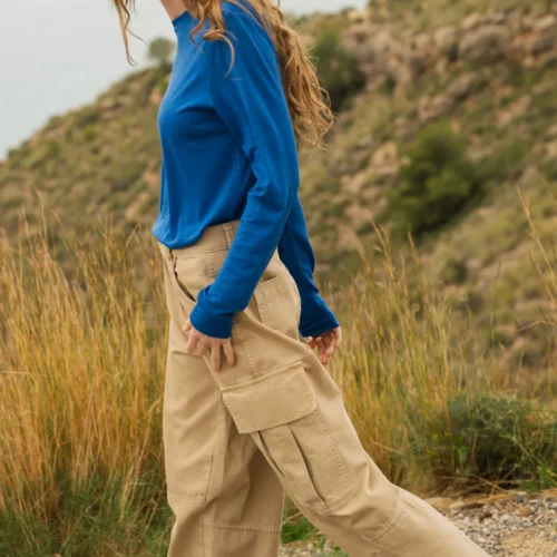 Osoba z długimi kręconymi włosami ubrana w niebieską bluzkę z długim rękawem i beżowe spodnie cargo idzie na zewnątrz żwirową ścieżką, z trawą i wzgórzem w tle.