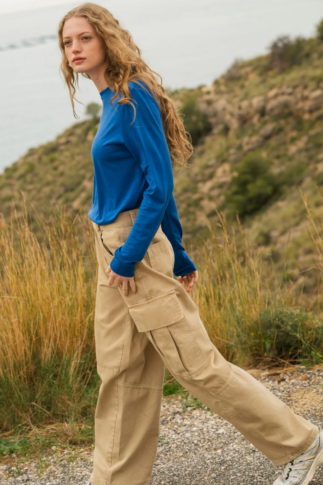 Osoba z długimi kręconymi włosami ubrana w niebieską bluzkę z długim rękawem i beżowe spodnie cargo idzie na zewnątrz żwirową ścieżką, z trawą i wzgórzem w tle.