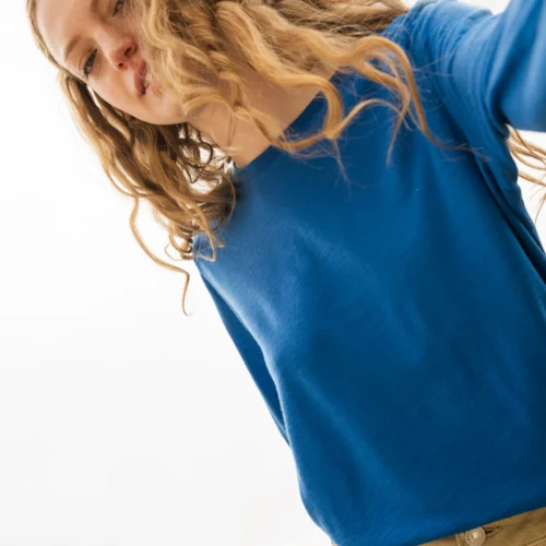 Osoba o długich, falowanych blond włosach ubrana w niebieską koszulę z długim rękawem i beżowe spodnie spogląda w dół na kamerę, z jasnym, zachmurzonym niebem w tle.