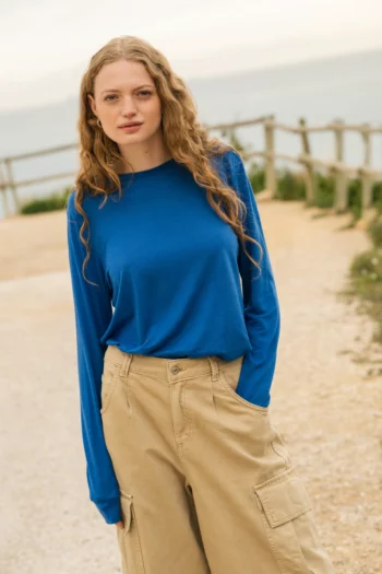 Młoda kobieta z długimi kręconymi włosami ma na sobie niebieską koszulę z długim rękawem i beżowe spodnie cargo, stojąc na zewnątrz na żwirowej ścieżce z drewnianym płotem i wodą w tle.