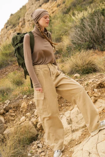 Osoba ubrana w beżową chustę na głowie, brązową koszulę z długim rękawem, luźne brązowe spodnie cargo i białe trampki wędruje na zewnątrz z dużym zielonym plecakiem po skalistym, trawiastym wzgórzu.