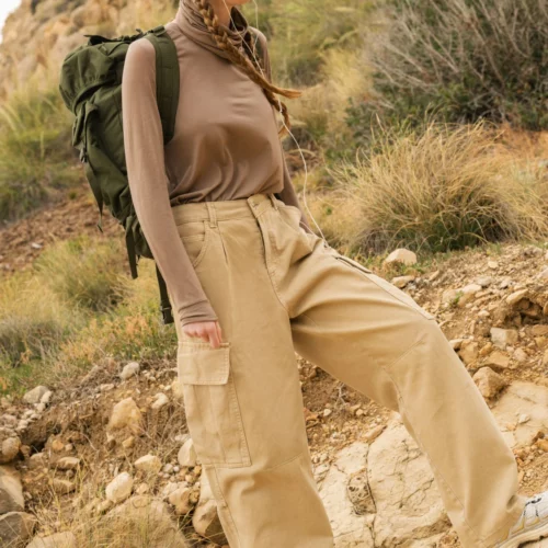 Osoba ubrana w beżową chustę na głowie, brązową koszulę z długim rękawem, luźne brązowe spodnie cargo i białe trampki wędruje na zewnątrz z dużym zielonym plecakiem po skalistym, trawiastym wzgórzu.