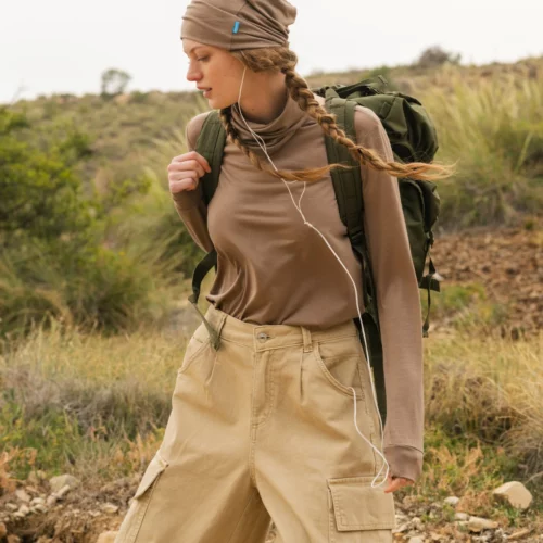 Kobieta ubrana w brązową koszulę z długim rękawem, spodnie cargo w kolorze khaki i pasującą czapkę idzie na zewnątrz z zielonym plecakiem i słuchawkami, otoczona skalistym terenem i trawiastymi wzgórzami.